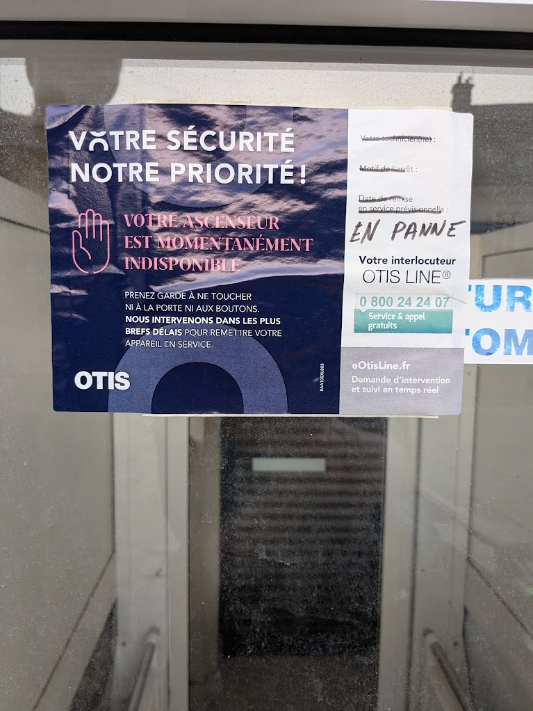 Une affichette Otis sur un élévateur. La mention « Date de remise en service prévisionnelle » est barrée et « EN PANNE » est écrit au feutre en dessous